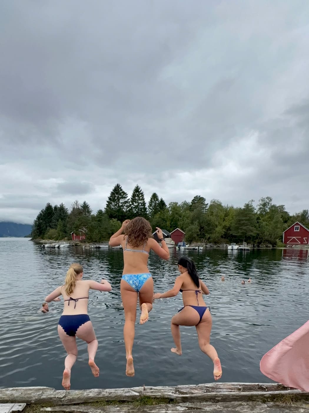 Three students wading into a fjord towards red boathouses on the far shore