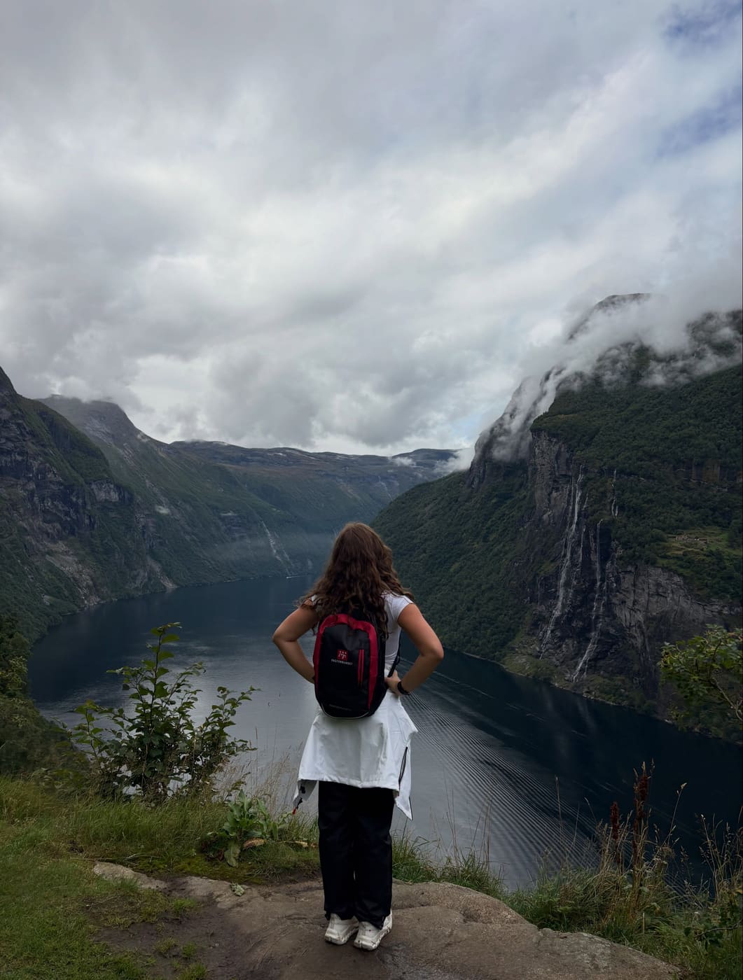 A student with a red backpack looking out over the Geirangerfjord, a waterfall on the far cliff