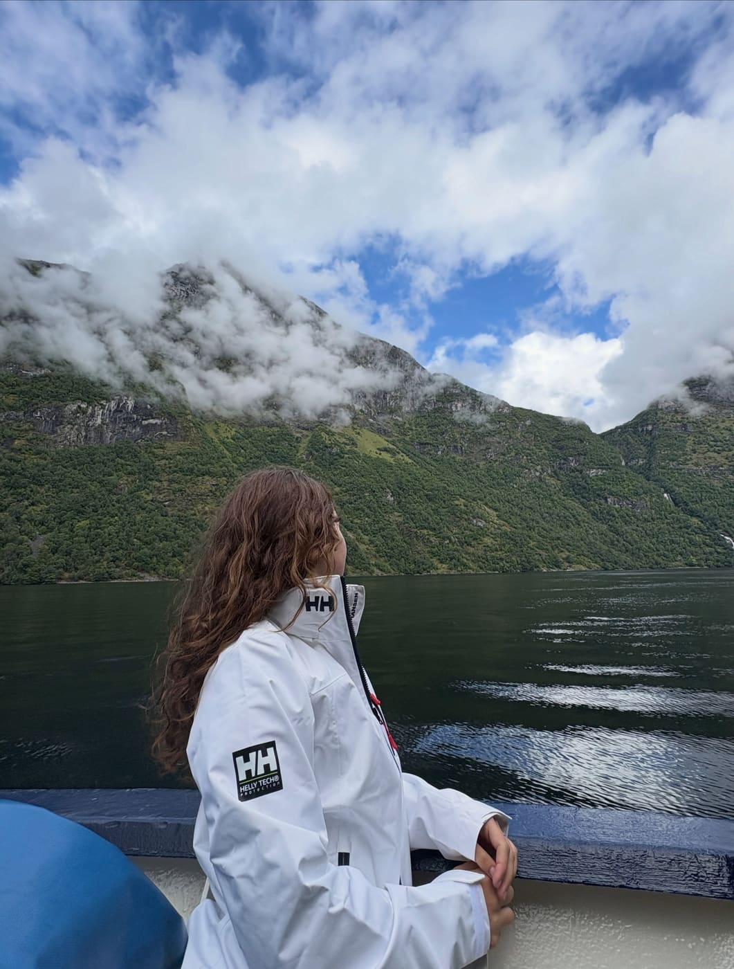 A student in a white jacket looking out across a fjord towards low cloud
