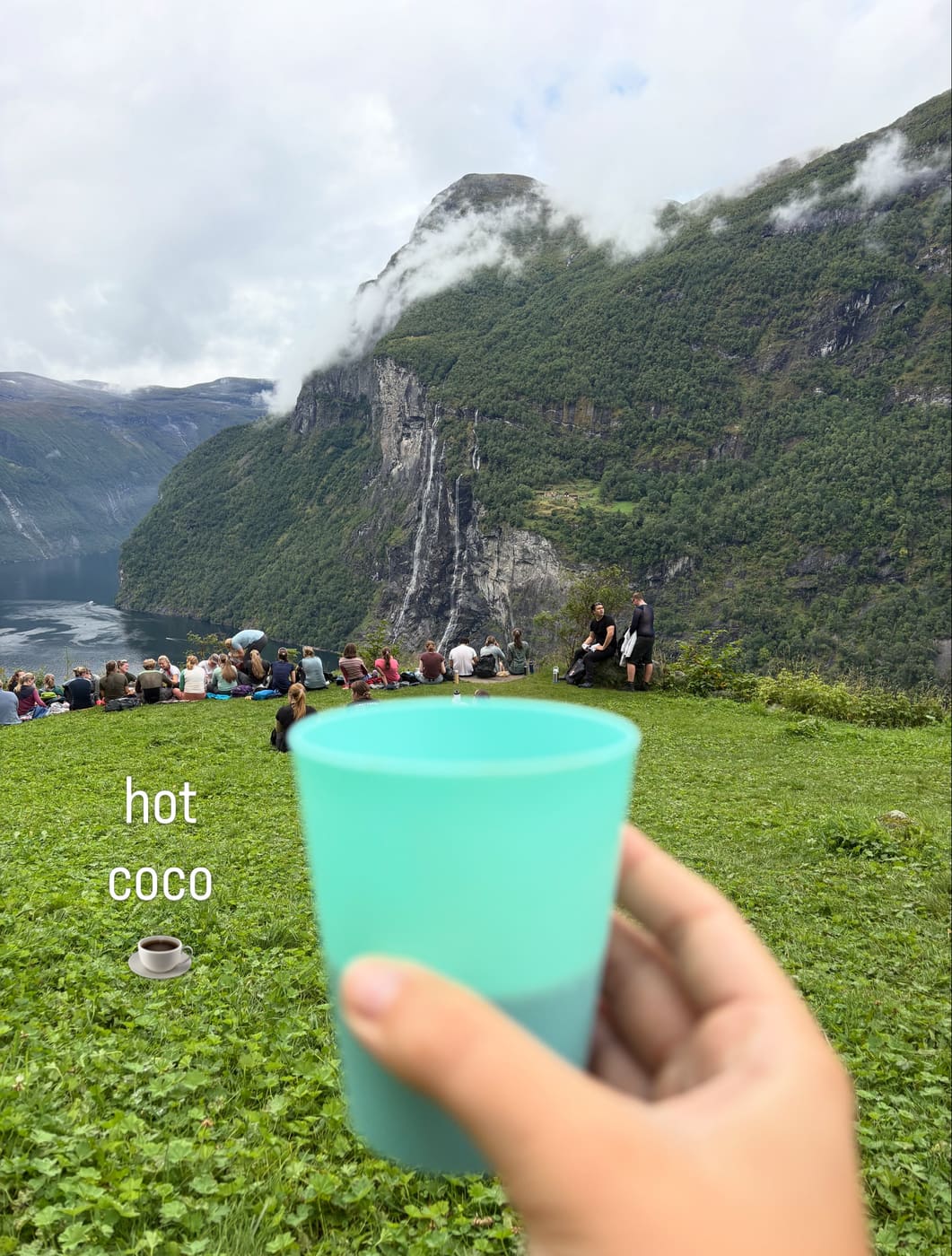 A group of students sitting on a grassy slope high above a fjord, the Seven Sisters waterfall opposite, one holding a cup of hot chocolate